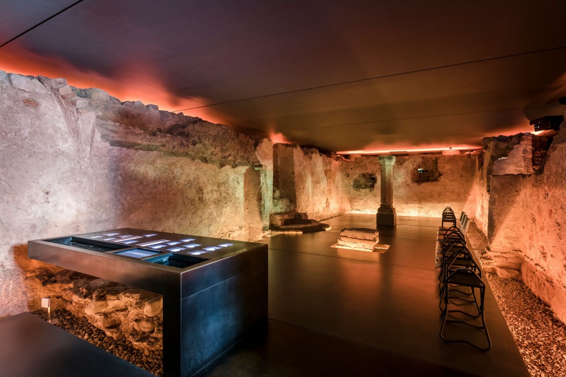 Unterirdischer Schauraum Erasmuskapelle mit altem Gemäuer. Im Raum stehen Stühle und ein Ausstellungstisch. Eine Multimediashow erweckt den Eindruck von brennenden Mauern..